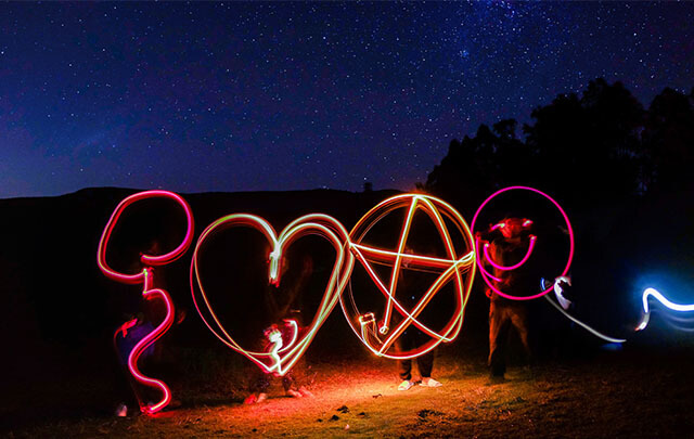 Four positive people standing in a field under a dark star lite sky using light to draw hearts, stars and smiling faces using slow exposure photography.