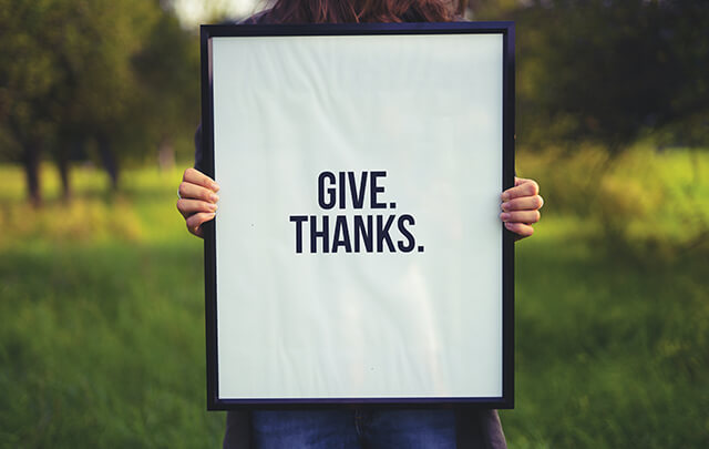 Woman standing in field holding sign that says Give Thanks.