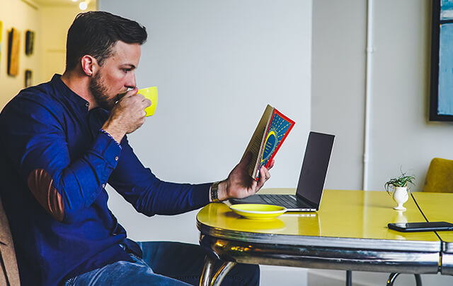 Man sitting at yellow table in sober living house, sipping out of a yellow mug reading a book.